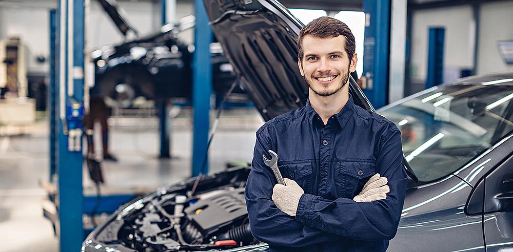 freundlicher junger Mechatroniker vor einem Fzg in der Werkstatt