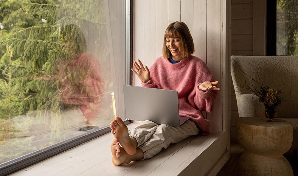 junge Frau sitzt gemütlich auf breiter Fensterbank an großem Fenster mit Sicht auf Wald und freut sich über etwas, das sie im Laptop sieht