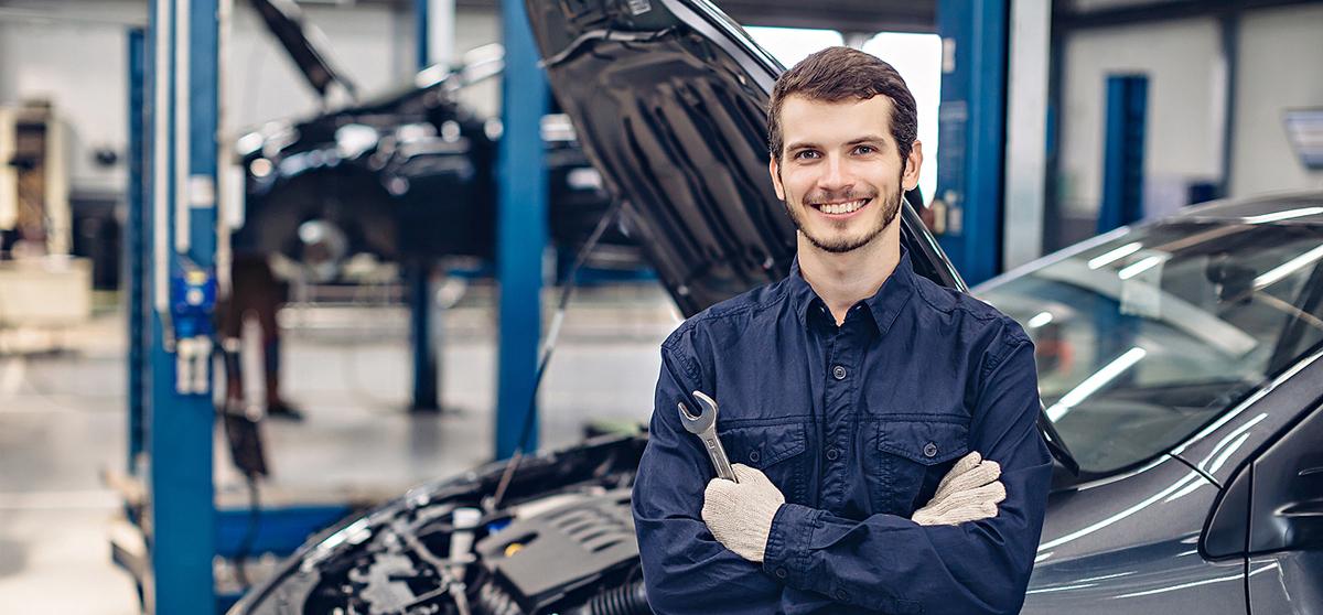 freundlicher junger Mechatroniker vor einem Fzg in der Werkstatt