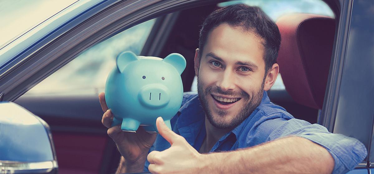 lächelnder junger Mann schaut aus Autofenster, zeigt freudig ein Sparschwein und zeigt "Daumen hoch"