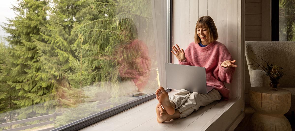 junge Frau sitzt gemütlich auf breiter Fensterbank an großem Fenster mit Sicht auf Wald und freut sich über etwas, das sie im Laptop sieht
