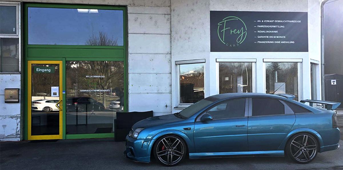Eingangsbereich der Firma Frey Cars mit großem Infoschild neben der Tür und einem geparkten Fahrzeug